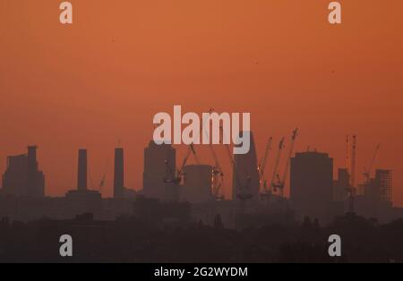 Wimbledon, London, UK. 13 June 2021. Orange and cloudless dawn sky begins a day of heat in London with temperatures due to reach 27 degrees by afternoon. Foreground cranes reflect  light from the rising sun with the four chimneys of former Battersea Power station in the distance. Credit: Malcolm Park/Alamy Live News. Stock Photo