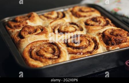 Freshly baked cinnamon rolls and cinnamon buns in a grey baking tin on top of a stove and on top of a table, drizzled with an icing sugar glaze. Stock Photo