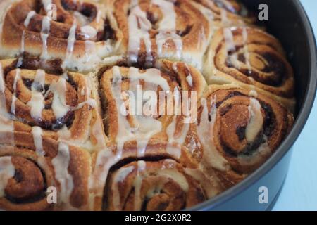 Freshly baked cinnamon rolls and cinnamon buns in a grey baking tin on top of a stove and on top of a table, drizzled with an icing sugar glaze. Stock Photo