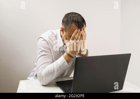 A hacker in handcuffs near a laptop, covers his face with his hands, a concept on the topic of computer crimes. Stock Photo