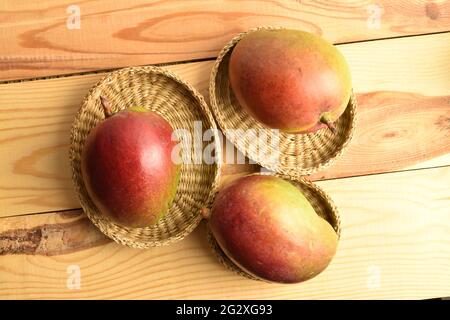 Three organic mangoes in straw plates, close-up, on a wooden table, top view. Stock Photo