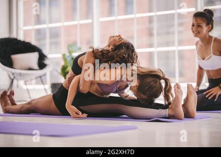 Two women doing partner exercise at yoga class with a friend looking on. Females doing workout at yoga class. Stock Photo