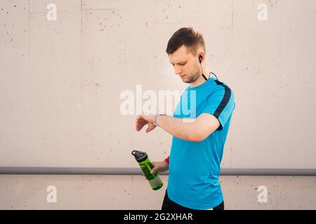 Man looks at smart watch, check the number of calories, heart rate, training time, in his hand a with a bottle of water, in summer in the city Stock Photo