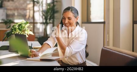 Woman executive sitting at desk talking on mobile phone. businesswoman speaking to client and making notes in coworking office space. Stock Photo
