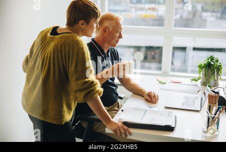Creative professionals working together on laptop. Two business people in office discussing work while looking at laptop, with man having coffee. Stock Photo