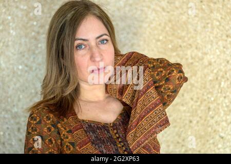 Attractive thoughtful woman holding up herarm as she looks wide eyed at the camera with a quiet pensive expression against a mottled wall Stock Photo