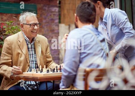 older caucasian male smiling, happy to win in chess game. sitting in outdoor cafe with his younger opponent who is talking to female. Stock Photo