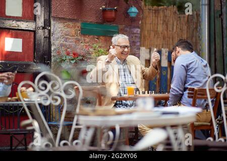 older caucasian male smiling with thumbs up. young guy with his hand over his eyes. sitting in outdoor cafe, playing chess. winning and loosing concep Stock Photo