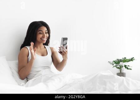 Hello. Positive black woman making video call via smartphone, waving hand while sitting on bed, empty space Stock Photo