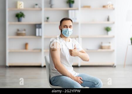 Young guy in face mask having plaster bandage on his arm after getting covid-19 vaccine injection, showing thumb up Stock Photo