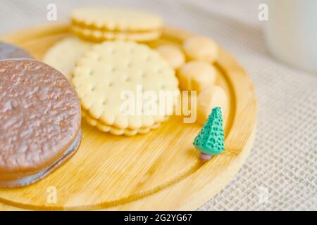 Cookies and cakes on a wooden tray. A small green Christmas tree made of plasticine. Chocolate sweets. The concept of Christmas and the New year. White cup on the background. Stock Photo