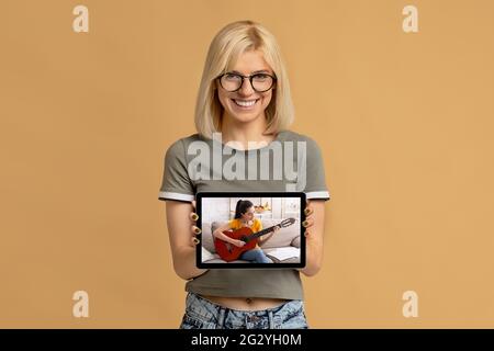 Young happy woman showing tablet pc with online guitar lesson on screen over beige studio background Stock Photo