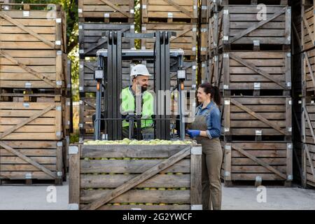 Happy young bearded guy driver in helmet in forklift truck with box full green apples Stock Photo