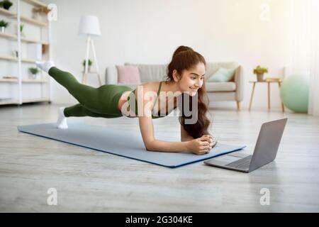 Strong Indian woman standing in elbow plank, lifting her leg on sports mat, following online video tutorial on laptop Stock Photo