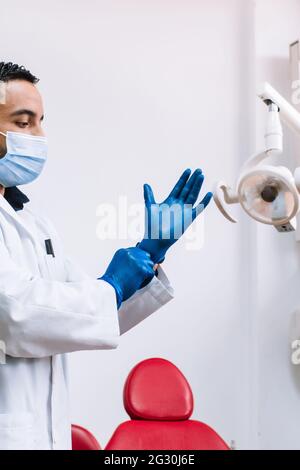 An adult dentist with a medical face mask putting on blue latex gloves Stock Photo