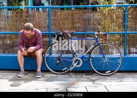 Man sits with his bicycle while looking at his phone, UK Stock Photo