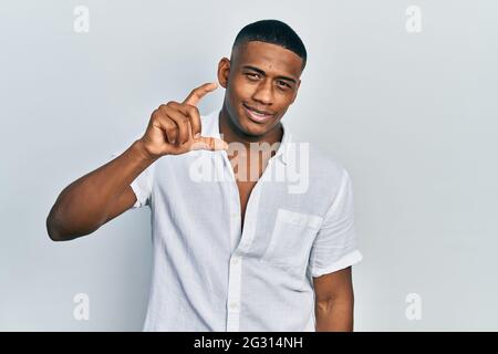 Young black man wearing casual white shirt smiling and confident gesturing with hand doing small size sign with fingers looking and the camera. measur Stock Photo