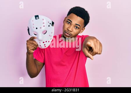 Young african american man holding hockey mask pointing with finger to the camera and to you, confident gesture looking serious Stock Photo