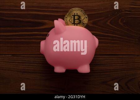 Happy cute pink plastic piggy bank with bitcoin coin inside on brown wooden planks background. Beautiful photo of a piggy bank and cryptocurrency Stock Photo