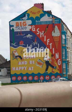 Sandylands Promenade, Heysham, Morecambe, Lancashire, United Kingdom. 9th June, 2021. Still hidden behind the scafolding the mural commissioned by MOrecambe’s Deco Publique and painted by Cobalt Collective with artists Erin Bradley-Scott and Kat Lowdon from the Cobalt Collective putting the finishing touch to the Shiftings Sands Mural which has been painted on to the gable of a house on Sandylands Promenade The Mural which is the work of the Glasgow based artis and Morecambes Deco Publique is a reminder to Morecambes glory days as a thriving seaside resort Credit: PN News/Alamy Live News Stock Photo