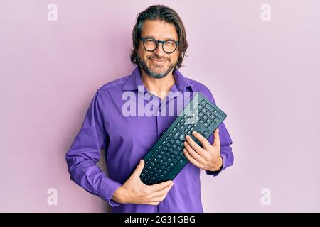 Middle age handsome man holding computer keyboard looking positive and happy standing and smiling with a confident smile showing teeth Stock Photo