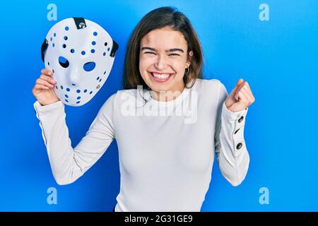 Young caucasian girl holding hockey mask screaming proud, celebrating victory and success very excited with raised arm Stock Photo