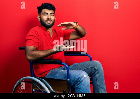 Arab man with beard sitting on wheelchair gesturing with hands showing big and large size sign, measure symbol. smiling looking at the camera. measuri Stock Photo