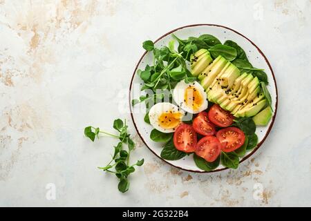 Avocado, cherry tomato, spinach and chicken egg, microgreens peas and black sesame seeds fresh salad in bowl on white stone table background. Healthy Stock Photo