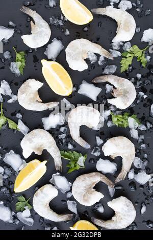 Seafood with lemon, parsley and ice on dark surface. Frozen raw shrimps. King Prawns on black stone background. Served food, preparing healthy food Stock Photo