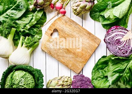 Fresh raw organic spring vegetables around cutting board top view on white wooden background. Vegan Genuine healthy eating concept. Stock Photo