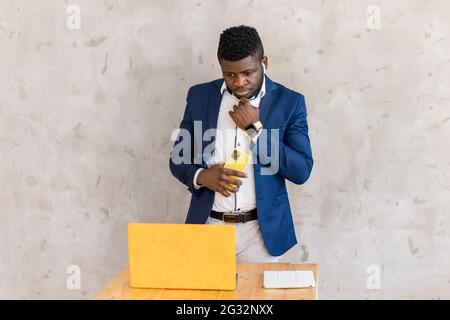 Pensive black working with yellow laptop at modern home or office. Focused african american businessman in blue suit, hold phone staying near desk and Stock Photo