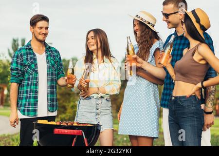 Young friends having fun grilling meat while enjoying barbecue, A group of happy guys and girls are preparing and eating on a barbecue dinner outdoors Stock Photo