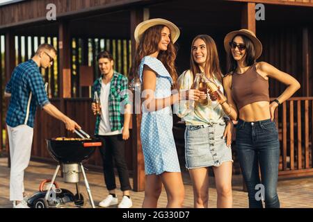 Young friends having fun grilling meat while enjoying barbecue, A group of happy guys and girls are preparing and eating on a barbecue dinner outdoors Stock Photo