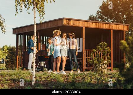 Young friends having fun grilling meat while enjoying barbecue, A group of happy guys and girls are preparing and eating on a barbecue dinner outdoors Stock Photo