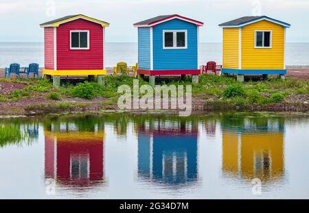 Three colorful small huts on a Newfoundland beach cast reflections on the water. Stock Photo