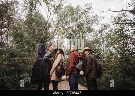 Four young people in warm clothes with guitar and backpacks standing in grove with green trees and looking back Stock Photo