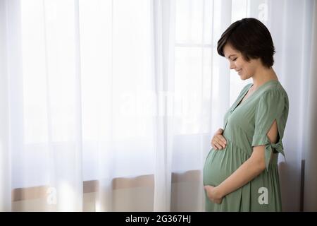 Side view of smiling pregnant woman embracing caressing belly