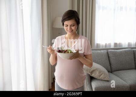 Smiling pregnant woman eating salad, fresh vegetables at home Stock Photo