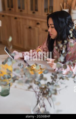 positive young asian woman in silk pajamas eating pancakes with fork and looking at cellphone with blank screen in kitchen Stock Photo