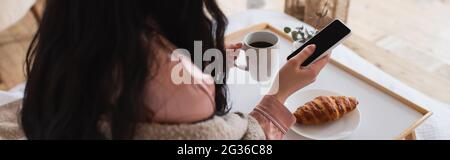 partial view of young brunette woman holding cellphone and having breakfast on bed, banner Stock Photo