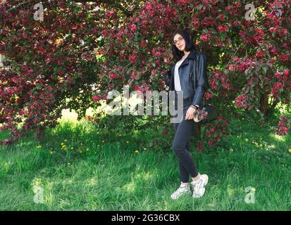 portrait of young beautiful brunette girl in black jacket and burgundy sweater standing near cherry blossoms on sunny spring day Stock Photo