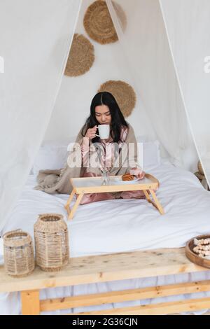 young asian woman in silk pajamas and blanket sitting on bed with crossed legs and drinking coffee in bedroom Stock Photo