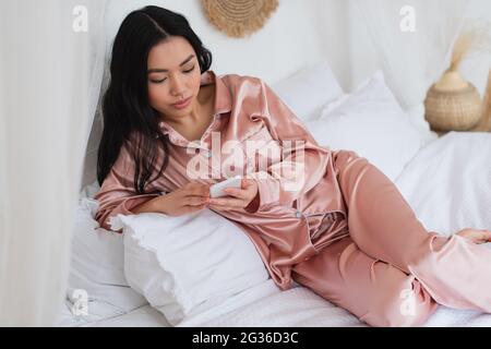 young asian woman in pink silk pajamas lying on bed and holding