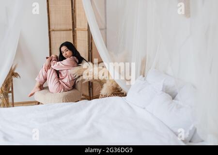 sad young asian woman sitting on chair in self hug pose and looking at camera in bedroom Stock Photo