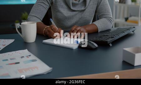Closeup of student with black skin writing communication homework on notebook while sitting at desk in living room. Young woman studying math on elearning platform doing homework during high school Stock Photo