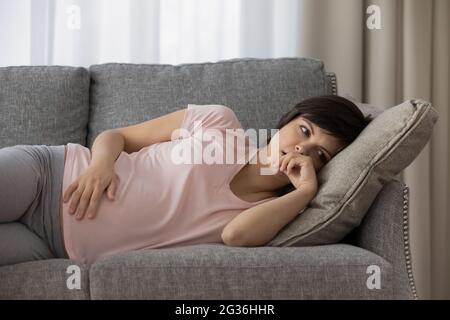Upset thoughtful pregnant woman lying on couch alone, touching belly Stock Photo