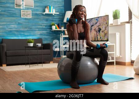 Smiling cheerful african woman flexing arm working out biceps, using dumbbells sitting on stability ball making training harder after warm up. Strong athletic person doing sports at home. Stock Photo
