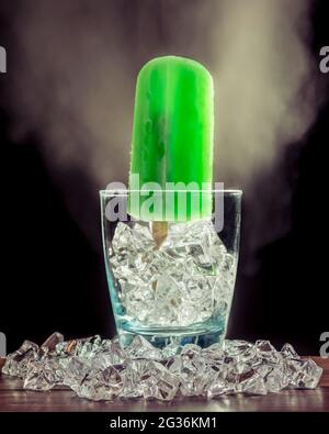 A green mint popsicle is standing inside a glass with ice and smoke on the dark background Stock Photo