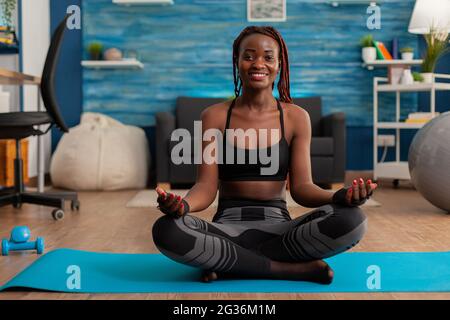 Fit black woman practicing yoga sitting with legs crossed in sportive top and leggins on lotus pose. Practicing calm mind harmony for no stress life in home living room. Stock Photo