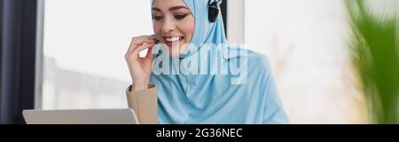 cheerful muslim woman adjusting headset while working in call center, banner Stock Photo